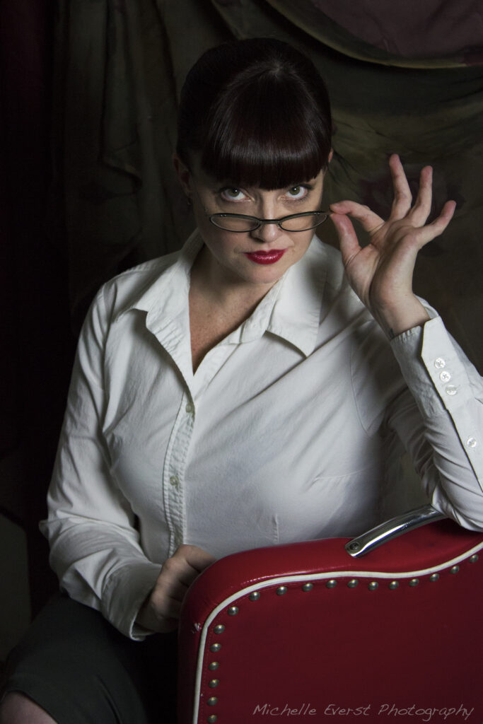 Kitten sitting in a red chair, wearing glasses and a white top.